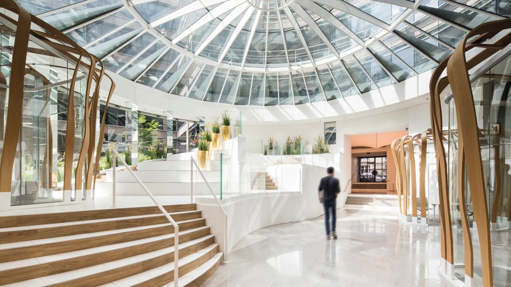 Moderne Eingangshalle mit Glasdach, geschwungenen Holzdetails, Treppen und Pflanzen, ein Mensch geht in der Mitte.