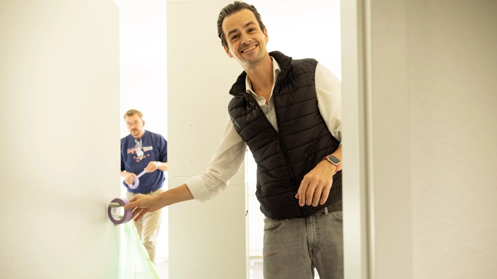 One man is holding tape, another is working on a wall in the background.