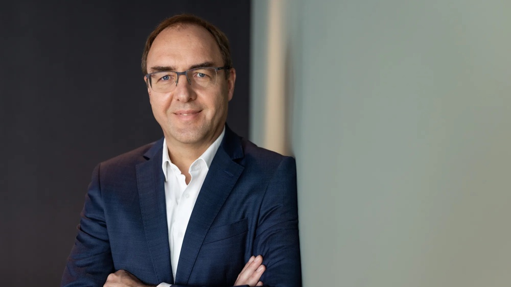 Vorstandsmitglied Dirk Schuster steht mit verschraenkten Armen vor einem dunklen Hintergrund