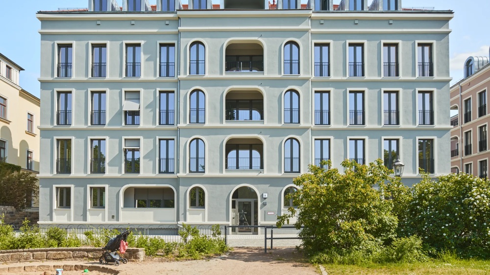 Fassade eines mehrstöckigen Gebäudes mit symmetrischen Fenstern und Balkonen, davor ein Kinderwagen und Büsche.