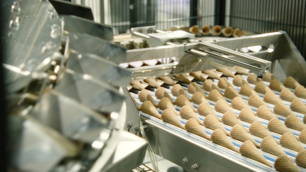 Zwei Fabrikationsmitarbeiter stehen in weißen Kitteln gebeugt über einer Eiswaffelproduktionsstraße und begutachten die Reihen der frischgebackenen Hörnchen.