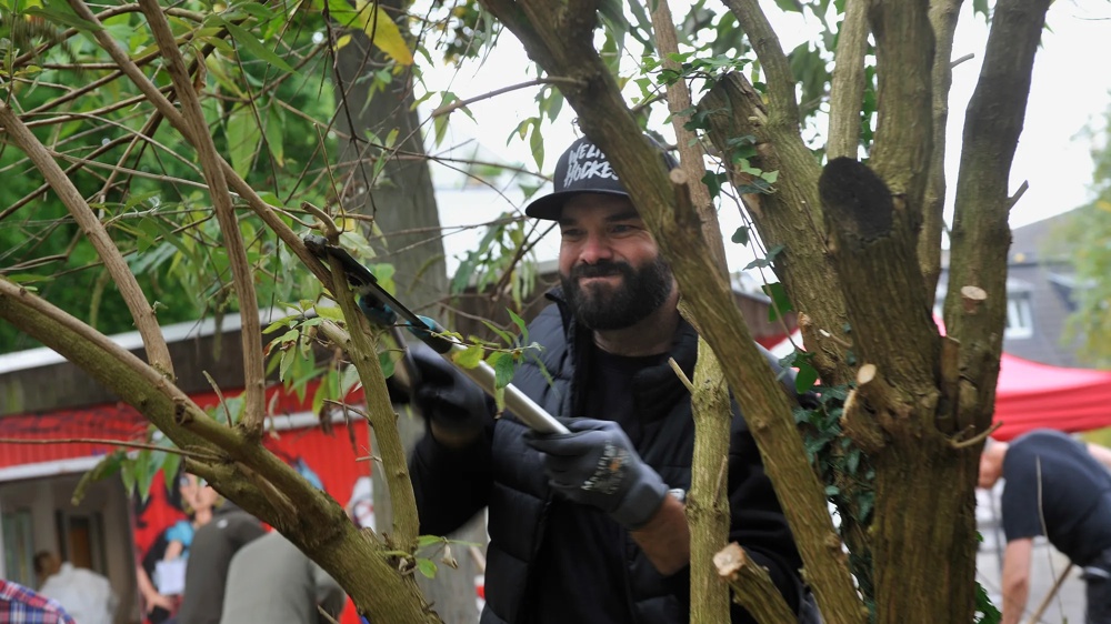 Mann mit schwarzer Mütze schneidet Äste eines Baumes im Freien, umgeben von anderen Menschen.