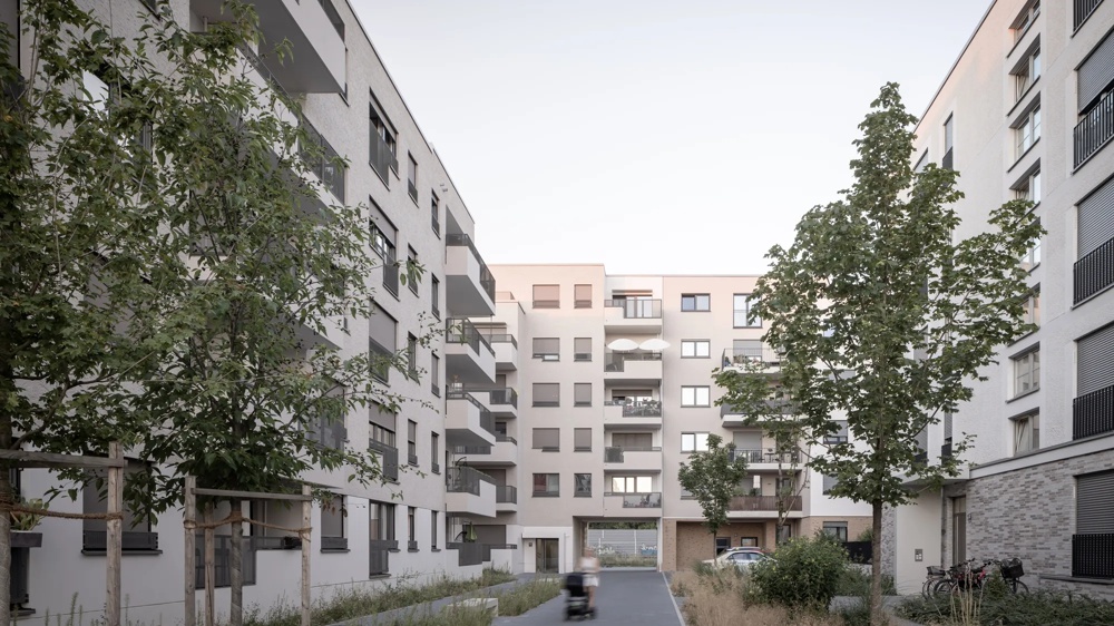 Wohnanlage mit Balkonen, Bäume im Vordergrund, Person im Rollstuhl auf Weg, Fahrräder rechts.