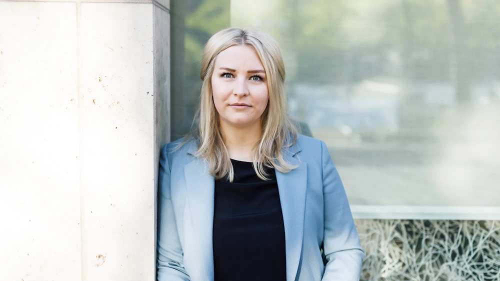 A woman in a blue blazer standing in front of a window looking into the camera.