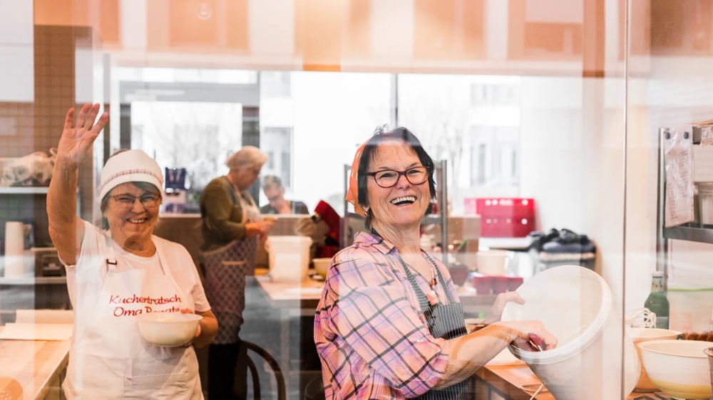 Zwei lachende Frauen in einer Bäckerei, eine winkt, die andere rührt Teig in einer Schüssel.