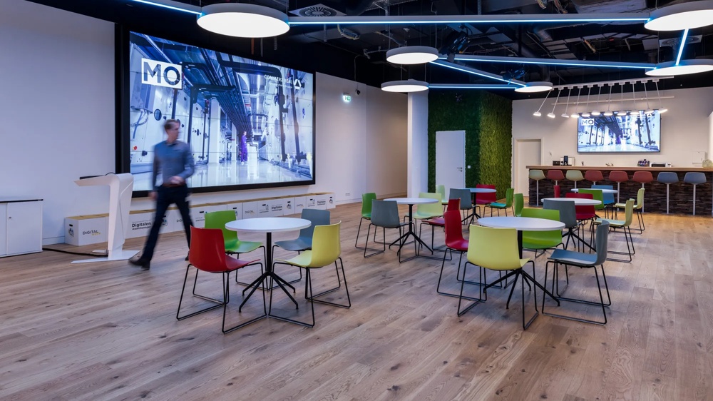 DigitalWerk. Com.Valley, event room. Machines are shown on the large screen and a large MO is on it. A man walking past the screen. Opposite the screen are table groups with colourful chairs.