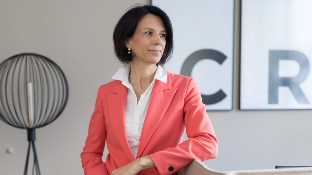 Frau in korallfarbenem Blazer lehnt an Tisch, moderne Lampe im Hintergrund.