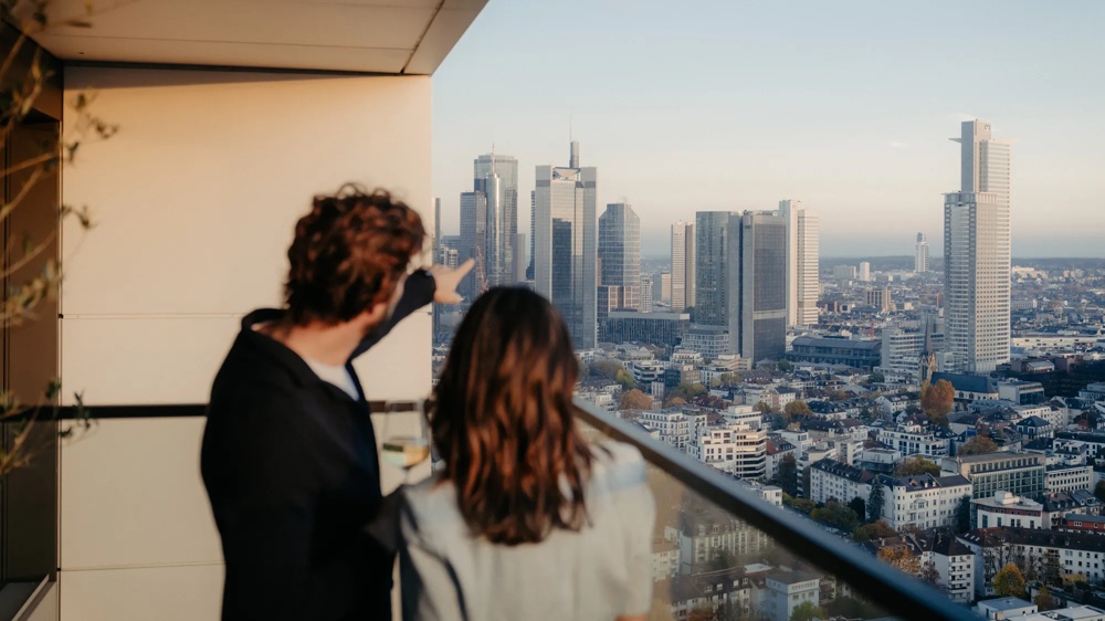 Zwei Personen auf Balkon einer modernen Wohnung mit Blick über die Stadt.