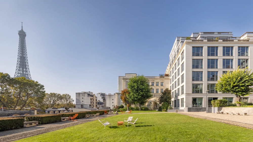 Grüne Wiese mit weißen Stühlen, modernem Gebäude und Eiffelturm im Hintergrund bei klarem Himmel.