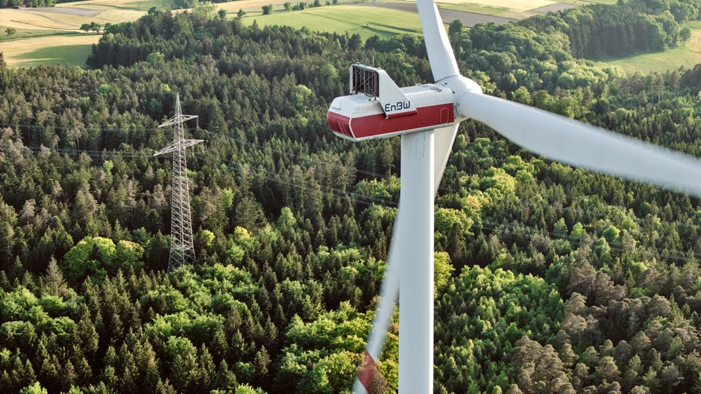 Drei Windräder stehen in einem grünen Wald unter blauem Himmel.