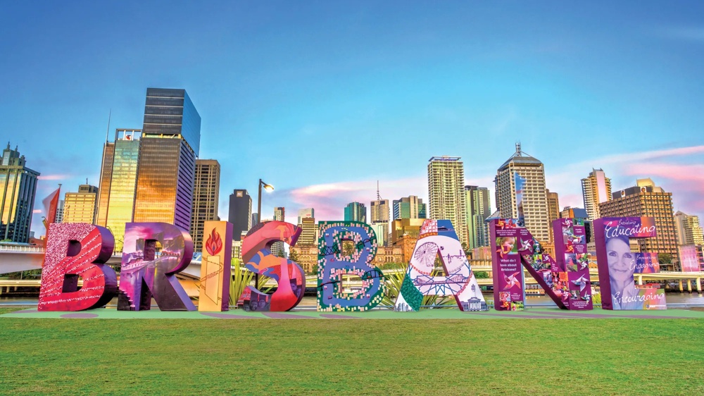 Bunte "BRISBANE"-Skulptur auf grünem Rasen vor einer Skyline bei klarem Himmel.