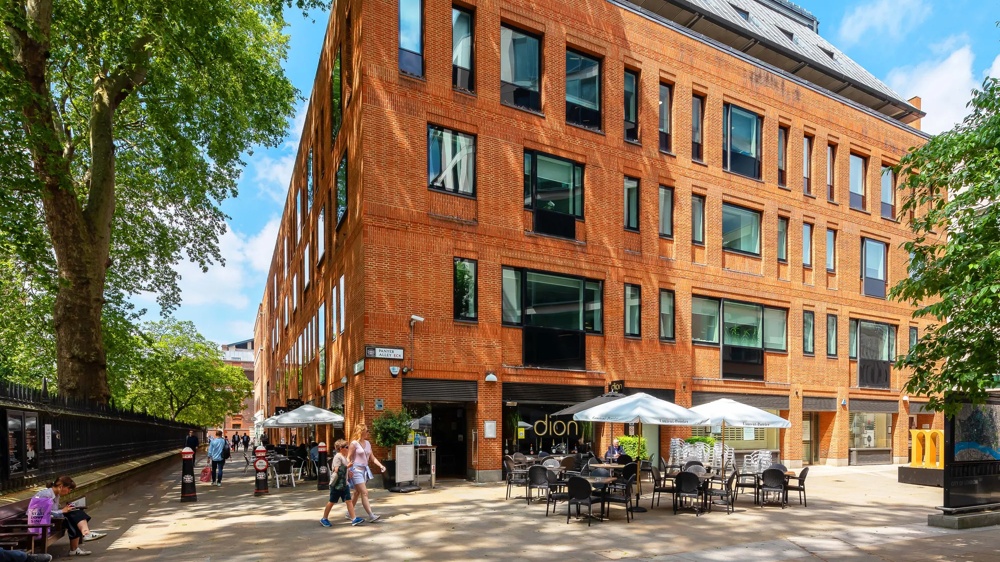 Red brick building with large windows, in front a café with white parasols, surrounded by trees.
