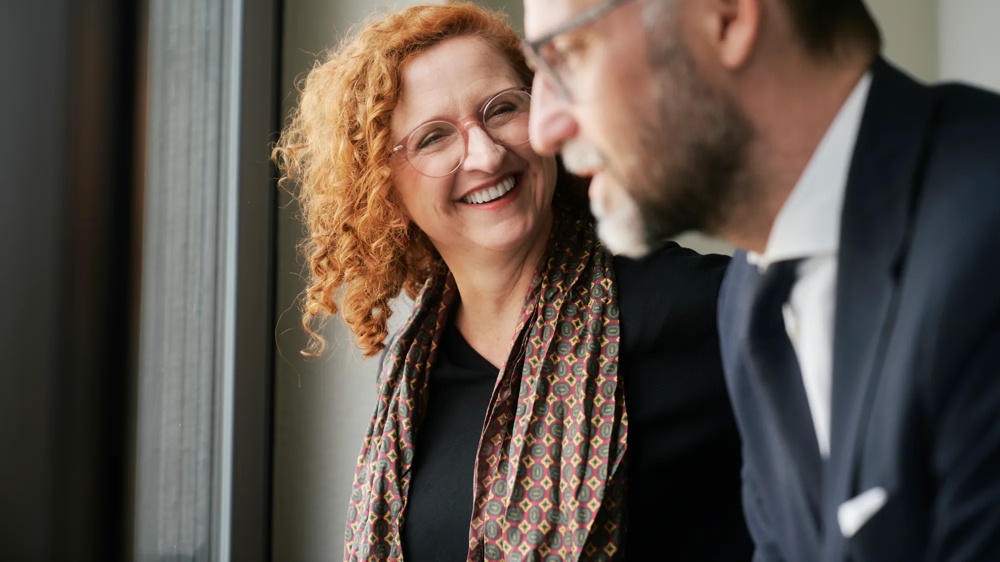 Eine lächelnde Frau mit lockigem Haar spricht mit einem Mann im Anzug vor einem Fenster.