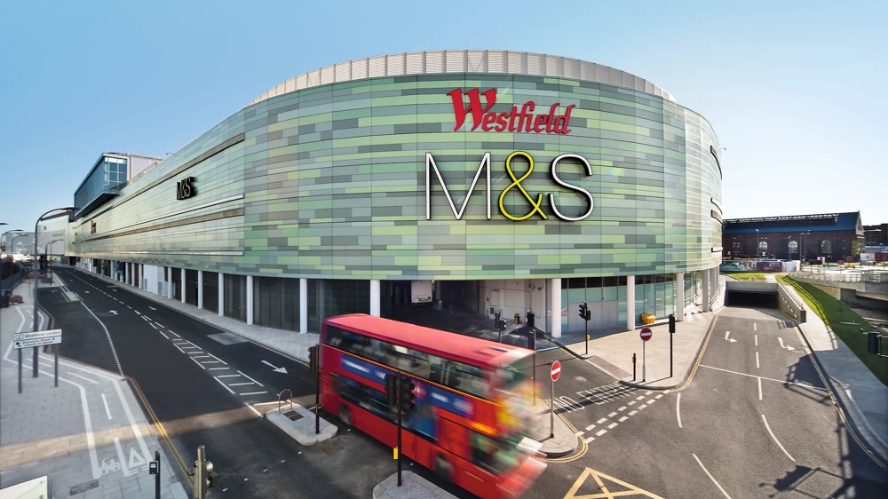 Grünes Einkaufszentrum mit M&S-Logo, roter Doppeldeckerbus auf Straße davor.