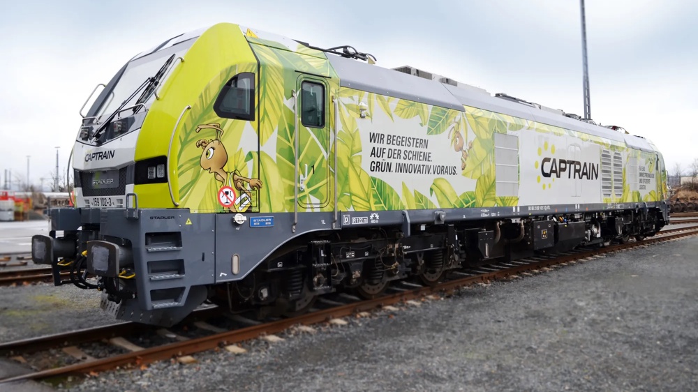 Grün-gelbe Lokomotive mit Blattdesign und Schriftzug "Wir begeistern auf der Schiene. Grün. Innovativ. Voraus." auf Gleisen.