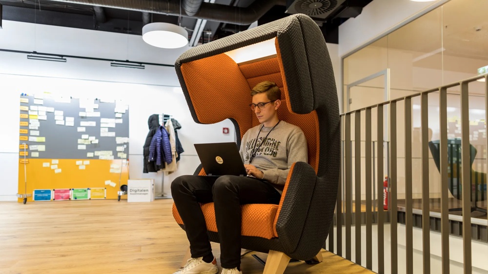 DigitalWerk A young man with glasses sitting in an orange ear chair looking at the laptop lying on his knees. In the background is a grey and orange mobile magnetic board with many white cards attached.