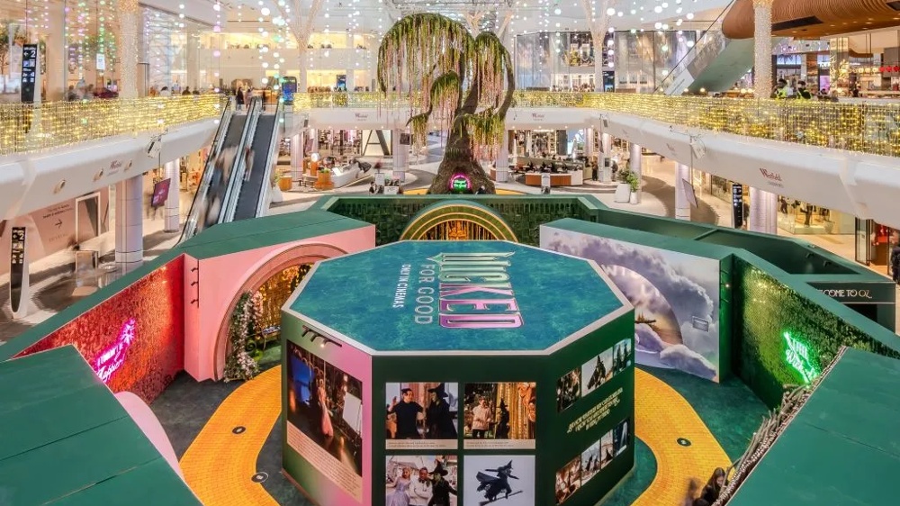 In Westfield London werden im Rahmen der Wicked: For Good Experience unter anderem Requisiten aus dem Film Wicked For Good ausgestellt. Auf dem Bild ist im Zentrum des Shopping Centers ein gruen-rosa Quader mit dem Wicked for Good Schriftzug auf seinem Dach zu sehen. An den Seiten befinden sich Plakate mit Bildern aus dem Film.  Um den Quader reihen sich Requisiten und Fotomotive aus dem Kinofilm. Um den Quader verlaeuft ein Pfad aus gelben Ziegelsteinen. Im Hintergrund sieht man das Atrium des Westfield London Shopping Centers.