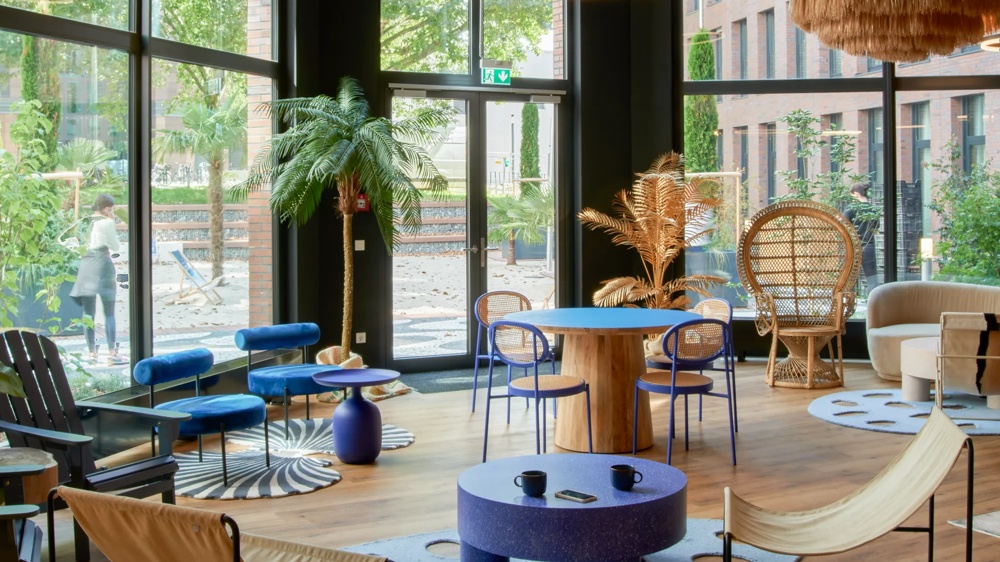 Modern room with blue furniture, wooden table, palm trees and large windows overlooking a green outdoor area.