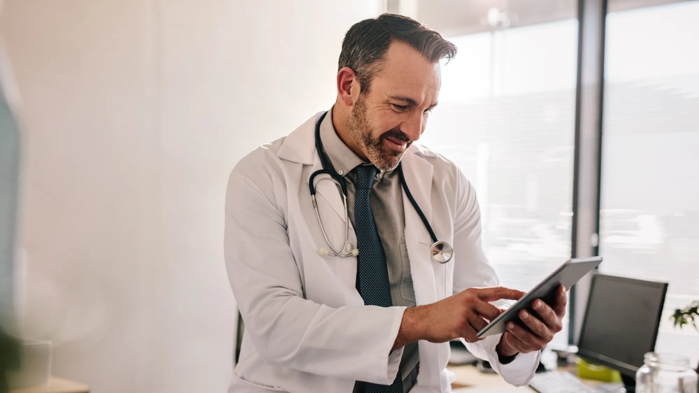 Ein Arzt im weißen Kittel benutzt ein Tablet in einem Büro neben einem Schreibtisch mit Computer.