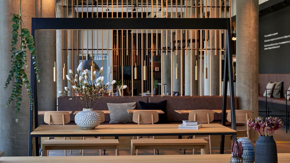 Moderner Raum mit Holztisch, Vase mit weißen Blumen, hängenden Lampen und Pflanzen im Hintergrund.