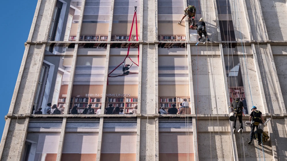 Am Berliner Alexanderplatz wird die Fassade der Galeria-Filiale gezeigt, wie sie von Fassadenkletterern mit bedruckten Klebebahnen versehen wird. die Bahnen visualisieren die mögliche neue Innengestaltung des Gebäudes als offene Bibliothek.