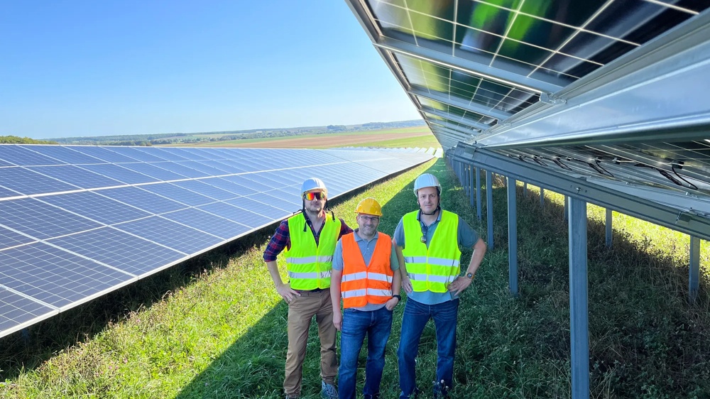 Drei Personen in Warnwesten stehen neben Solarpaneelen auf einem Feld unter klarem Himmel.