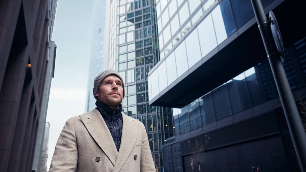 Ein Mann in beigem Mantel und Mütze steht vor modernen Hochhäusern in der Stadt.