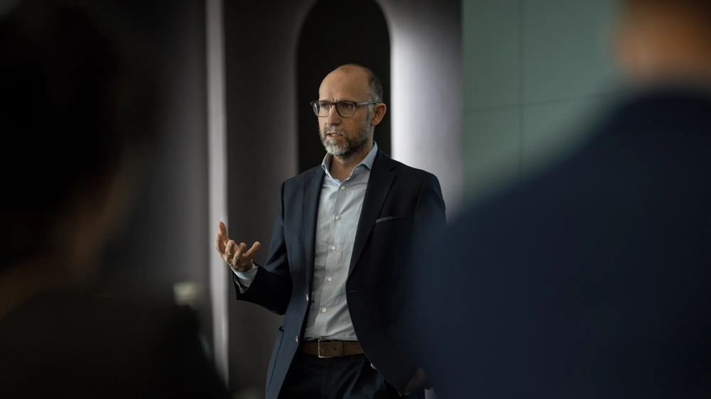 Ein Mann in Anzug spricht gestikulierend vor einem unscharfen Hintergrund.