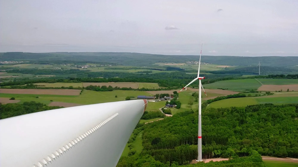 A close-up of a wind turbine wing over a green landscape with a wind turbine in the background.
