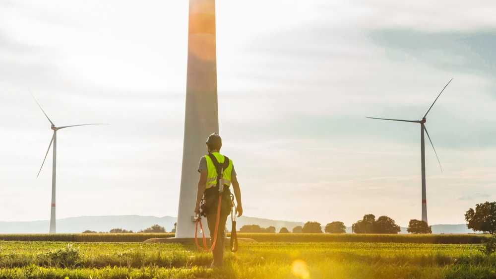 Ein Arbeiter in Schutzkleidung steht vor drei Windkraftanlagen auf einem Feld bei Sonnenuntergang.