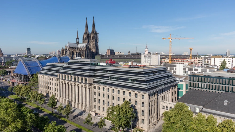 Historische Gebäude mit Kölner Dom im Hintergrund, blauer Himmel, Bäume und Baukräne.