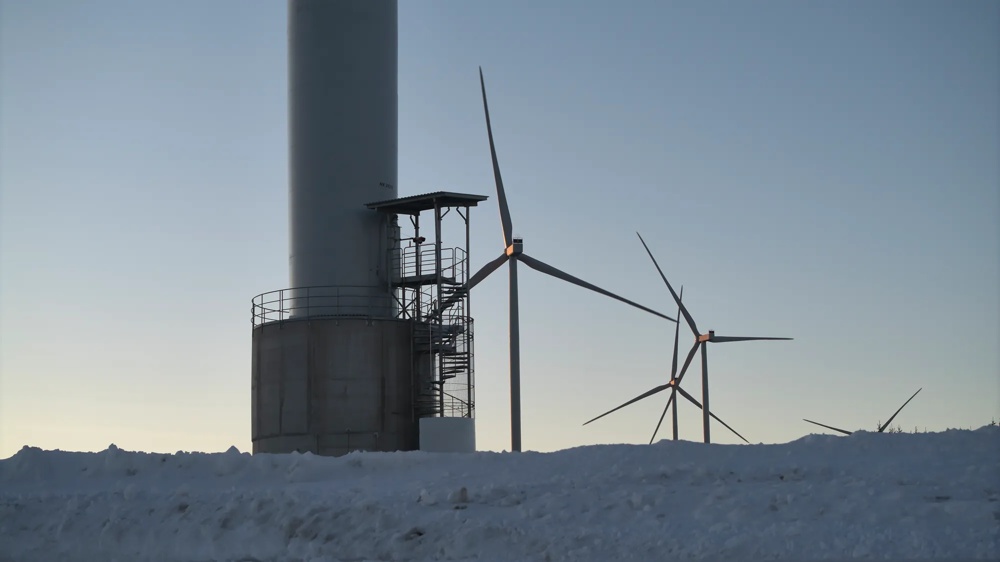 Im Schnee steht der Sockel eines Windrads mit Treppe. Im Hintergrund stehen weitere Windräder