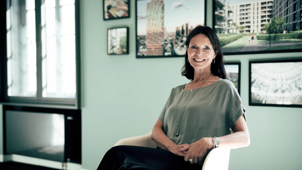 Frau sitzt lächelnd auf Stuhl vor mintgrüner Wand mit gerahmten Bildern.