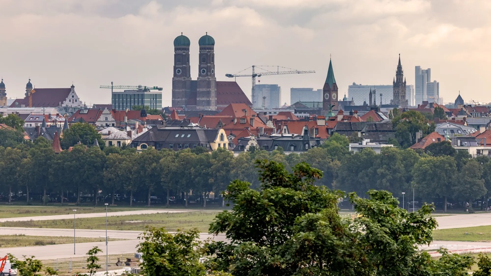 Stadtansicht mit Kirchtürmen, Baukränen und Wolkenkratzern im Hintergrund, Bäume im Vordergrund.