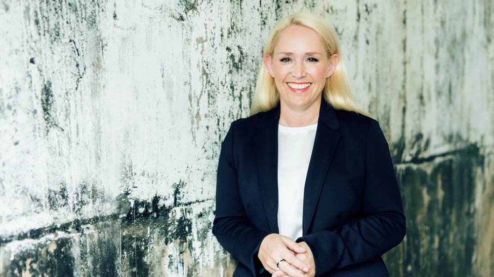 Woman in dark suit smiling in front of a structured, weathered concrete wall.