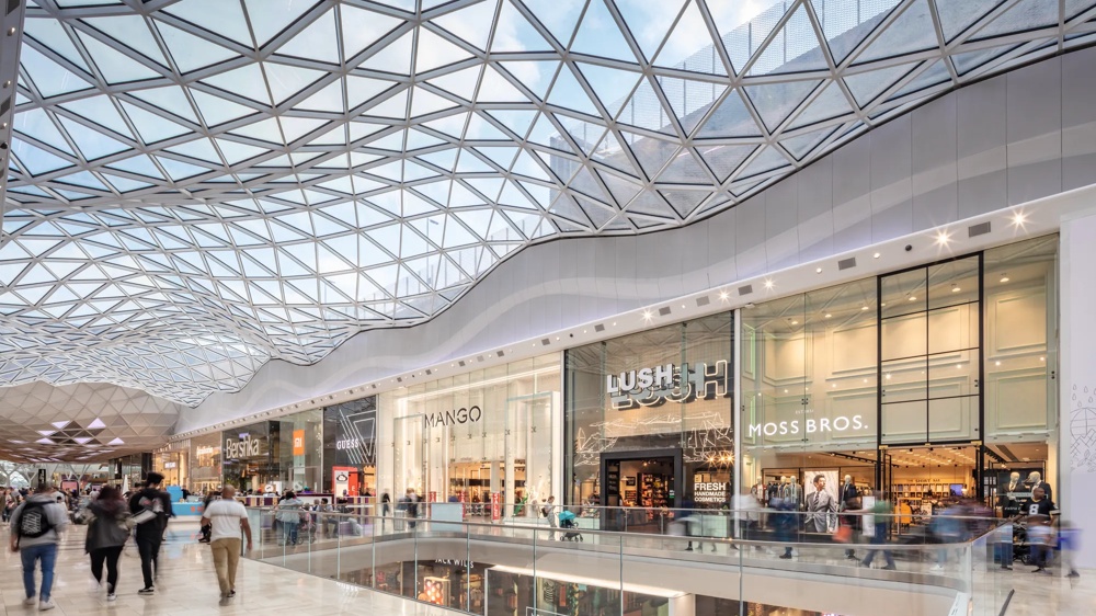 A modern shopping mall with a glass roof, shops and people strolling along a railing.