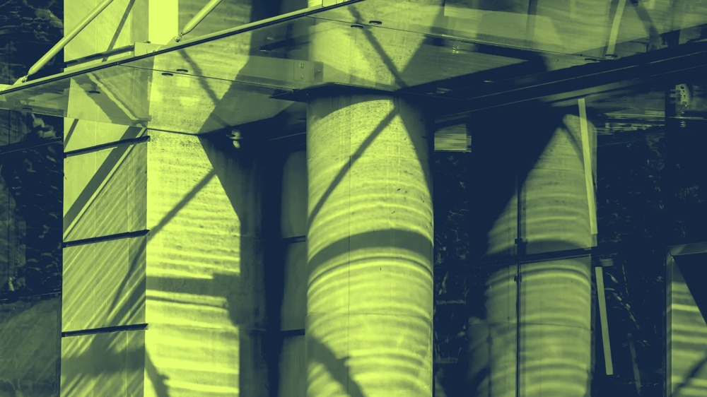 Schattenmuster auf Glasfassade mit Betonpfeilern und Metallstrukturen in gelbgrünem Licht.