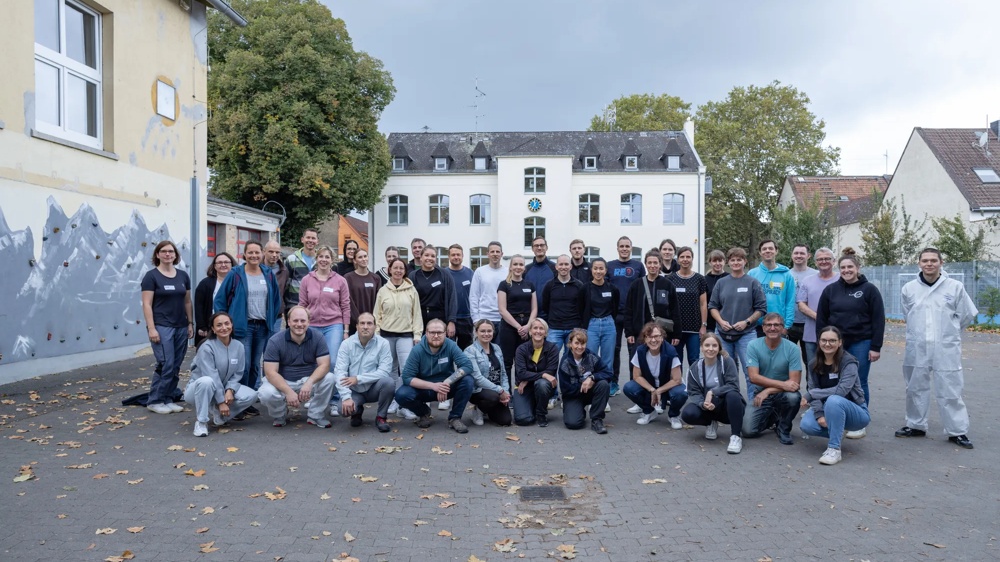 Eine Gruppe von Menschen posiert draußen vor einem Gebäude mit Bäumen im Hintergrund.
