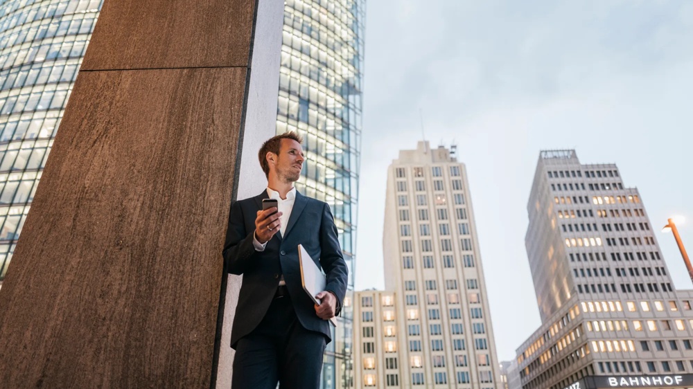 Ein Mann im Anzug steht mit Handy und Laptop vor hohen Bürogebäuden.