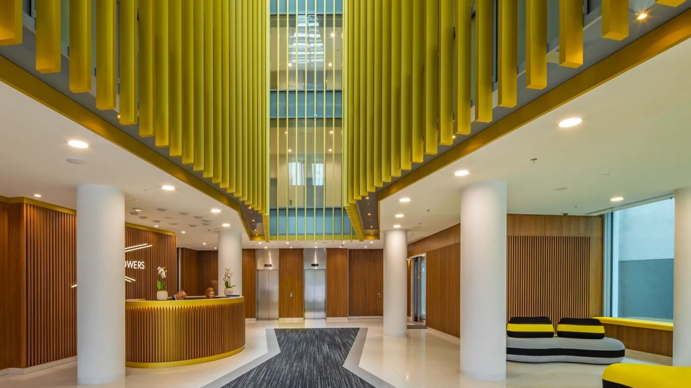 Empfangsbereich mit Holztheke, gelben Lamellen, großem Teppich, Sofas und Aufzügen im Hintergrund.