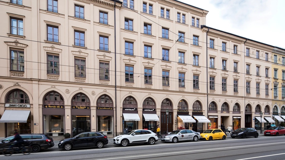 Elegant, multi-storey building with arcades and shop windows, cars and passers-by parked in front.