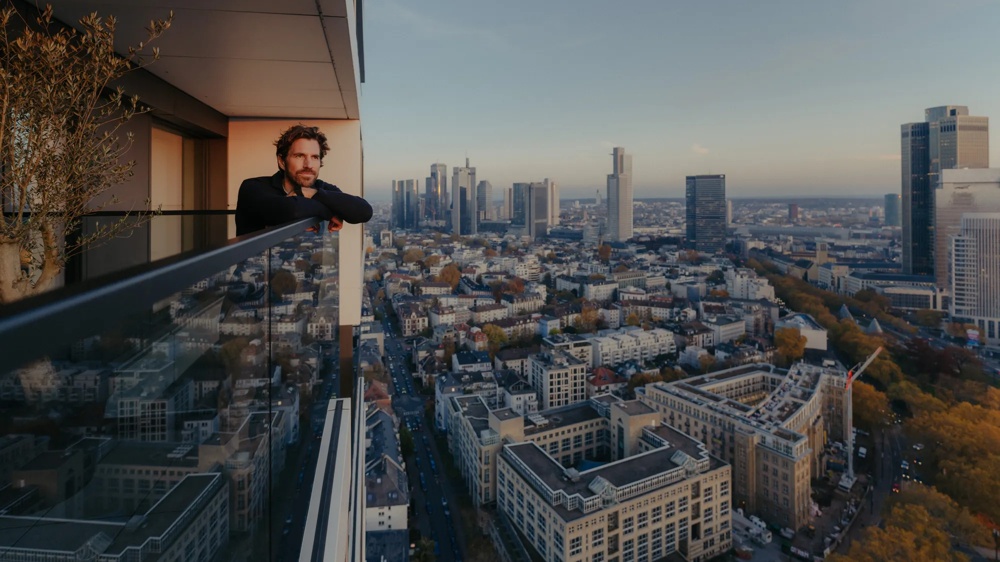 Person auf Balkon einer modernen Wohnung mit Blick über die Stadt und Getränk in der Hand.