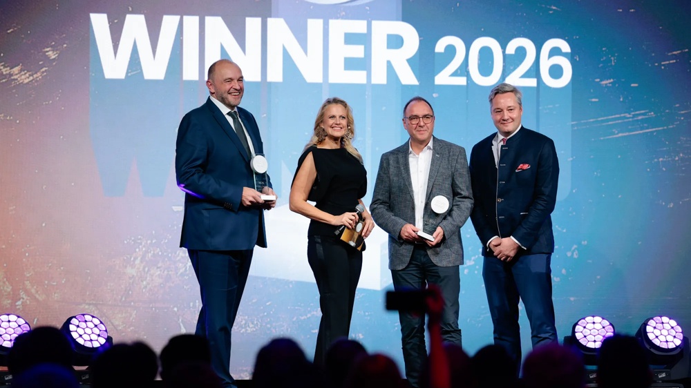 Vier Personen stehen auf einer Bühne mit Trophäen, vor einem Hintergrund mit "WINNER 2026".