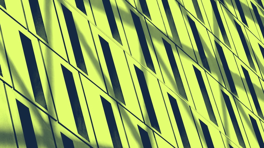 Gelbe Wand mit diagonalen, schmalen, dunklen Fenstern im geometrischen Muster, Schatten fallen schräg über die Oberfläche.