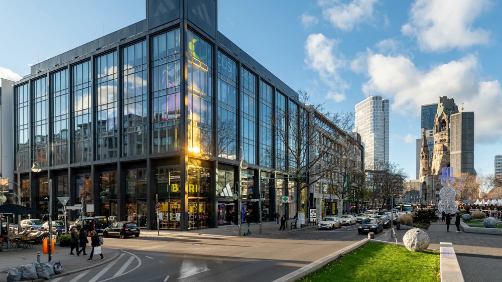 Moderne Glasfassade eines Gebäudes bei Sonnenschein, Menschen und Autos auf der Straße, blauer Himmel mit Wolken.