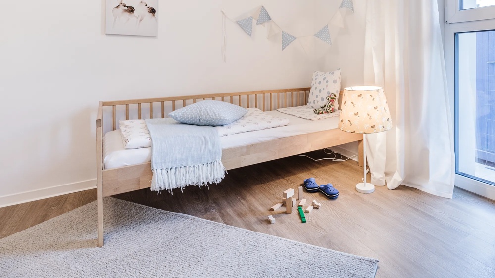 Holzbett mit blauer Decke, Kissen, Lampe, Spielzeug und Fenstern in hellem Kinderzimmer.