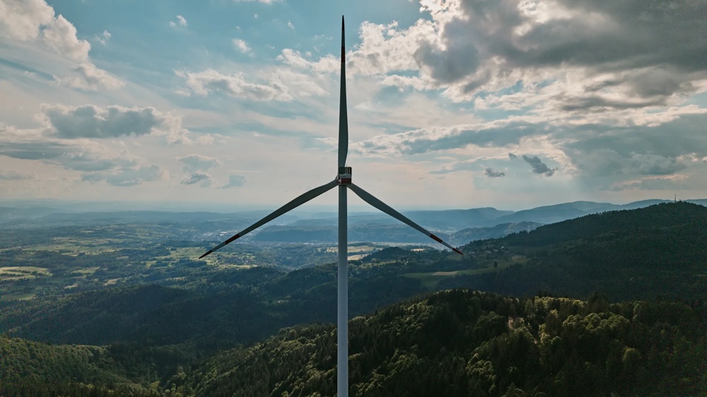 Zwei Windräder stehen auf einem bewaldeten Hügel unter wolkigem Himmel, umgeben von grünen Feldern und Bergen.