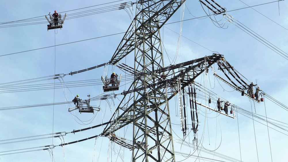 Arbeiter auf Hebebühnen reparieren Stromleitungen an einem großen Strommast unter blauem Himmel.
