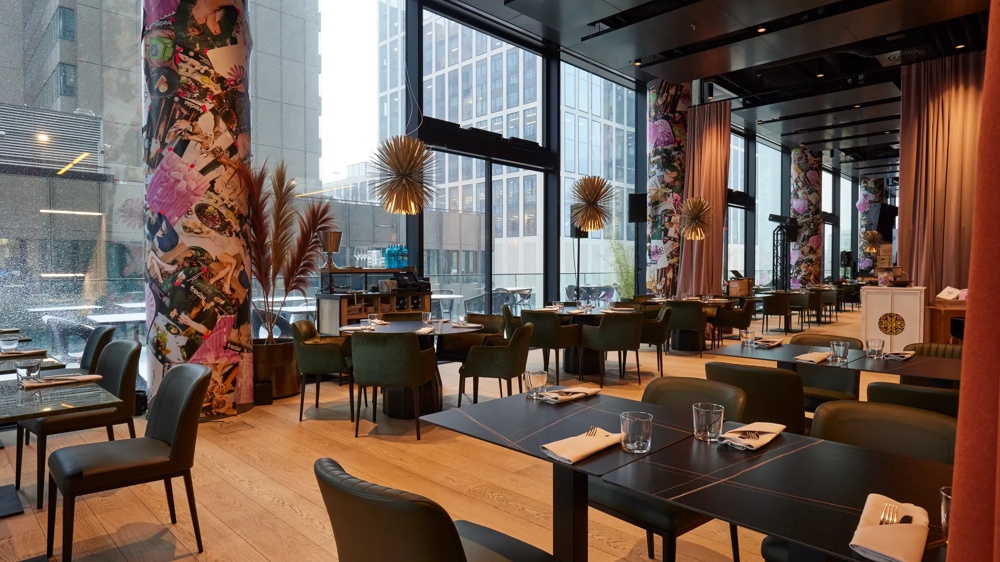 Modernes Restaurant mit großen Fenstern, dekorierten Säulen, grünen Stühlen und gedeckten Tischen auf Holzboden.