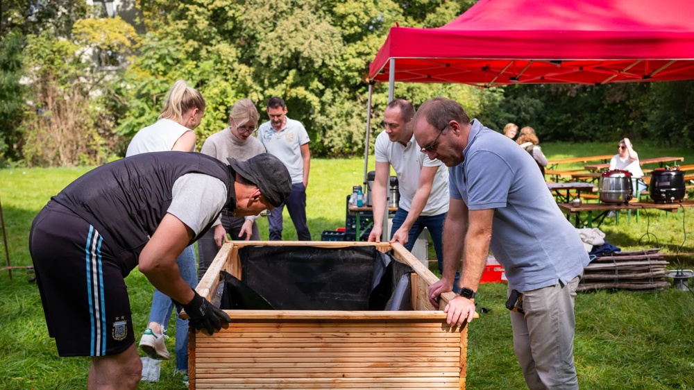 People together build a wooden planter under a red tent outdoors.